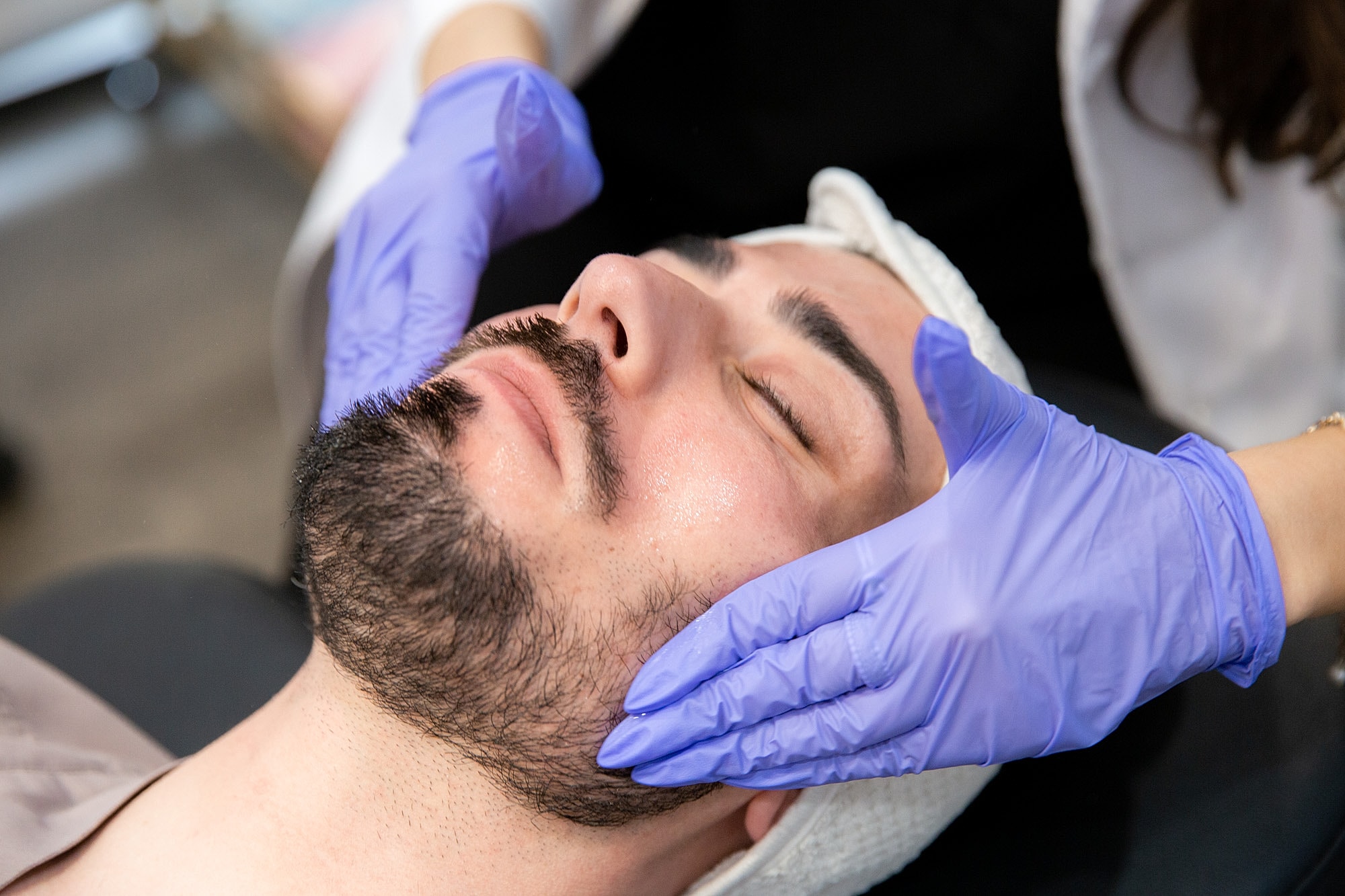 Man receiving facial treatment with gloved hands.